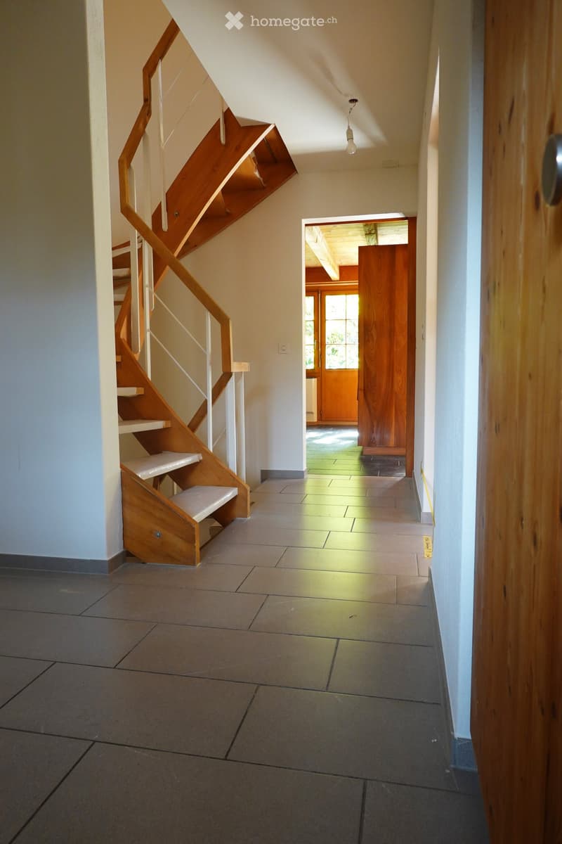 A modern hallway with a wooden staircase on the left and a doorway leading to another room on the right. The floor is tiled, and the walls are painted in a light color.