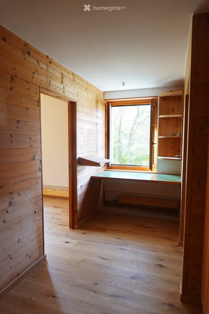 Empty room with wooden walls and floor, large window with open shutters, small built-in shelf, and a small desk area.