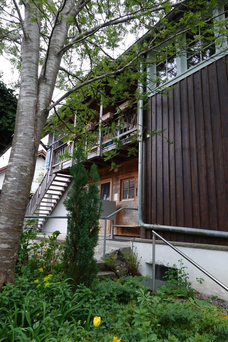 A two-story wooden house with a raised ground floor, featuring a staircase leading to an upper balcony, surrounded by greenery and flowers.