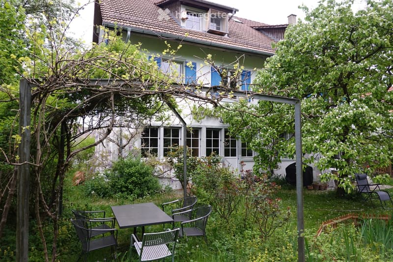 Garden area with a table and chairs, surrounded by greenery and trees, with a house in the background featuring multiple windows and a tiled roof.