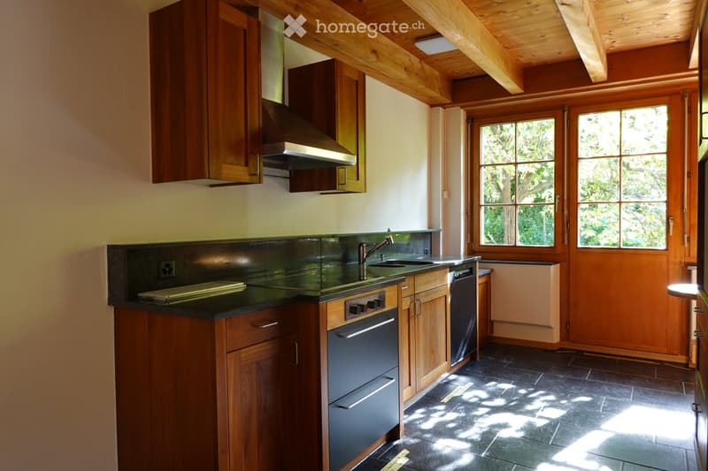 Bright kitchen with wooden cabinets, stainless steel appliances, and a dark countertop. Large windows allow natural light to fill the space.
