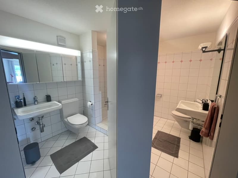 Two adjacent bathrooms with white tiled walls and floors, each equipped with a sink, toilet, and mirror. One bathroom features a dark-colored bath mat and a trash bin, while the other has a brown towel and a small trash bin.