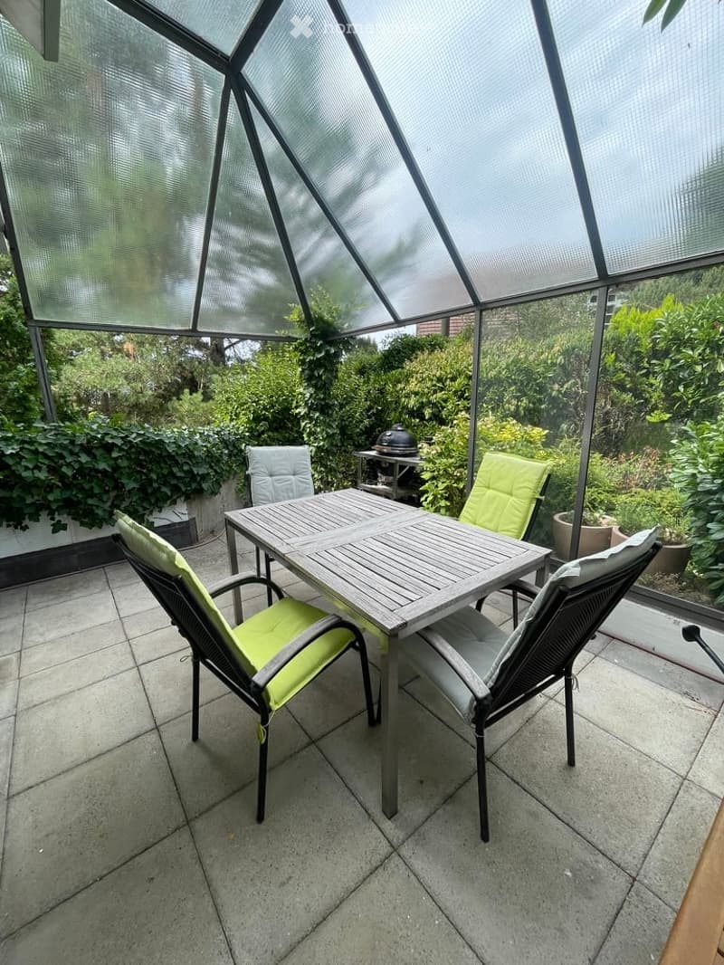 A modern glass-enclosed patio with a wooden dining table and four green-cushioned chairs, surrounded by lush greenery and a tiled floor.