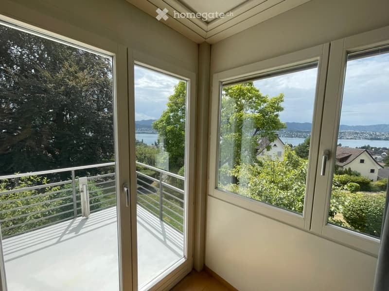 Balcony with metal railing, large windows offering scenic views of trees, water, and distant mountains.