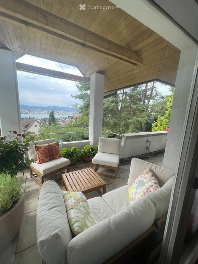 Balcony with wooden decking, white furniture including sofa, chairs, and coffee table, potted plants, scenic view of water and mountains.