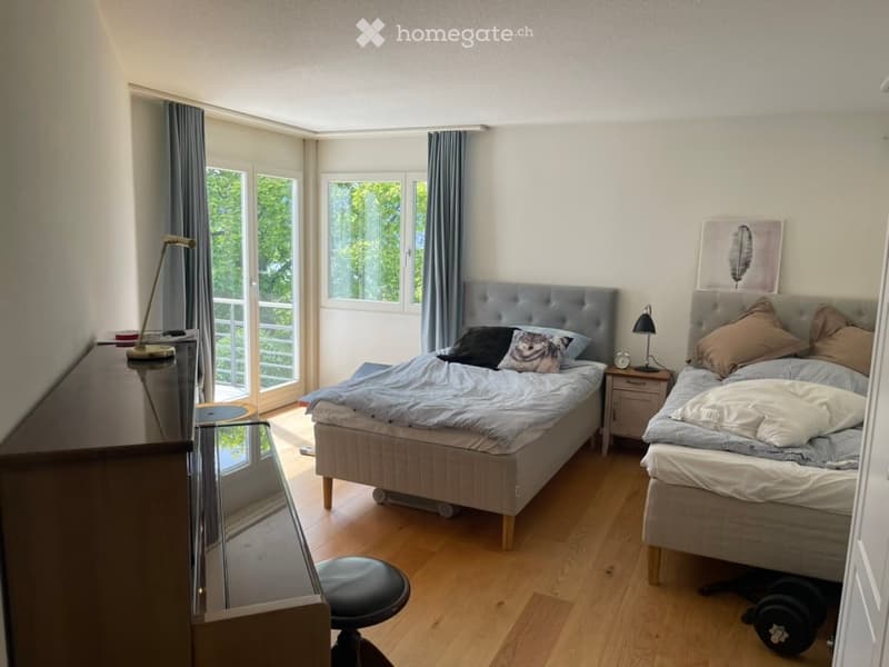 Bright bedroom with two single beds, nightstands, a stool, and a mirror. Large windows and a balcony door provide natural light and access to the balcony.