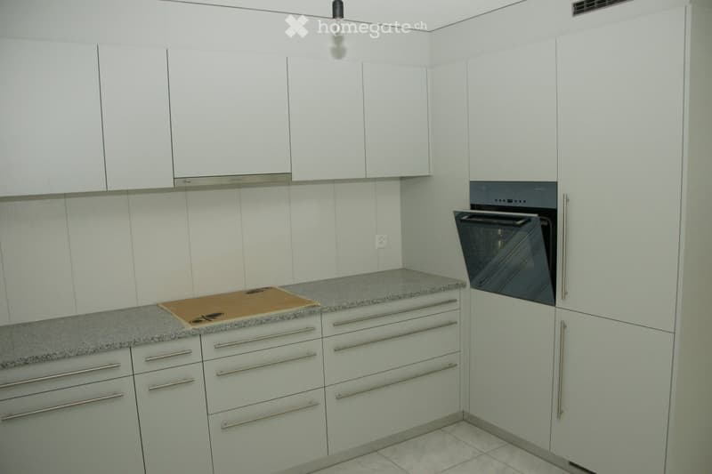 Modern kitchen with white cabinets, stainless steel handles, and a grey countertop. An oven is integrated into the cabinetry, and a cutting board rests on the counter.