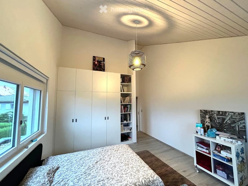 Bright bedroom with a large window, a neatly made bed, built-in white wardrobe, bookshelf, small table with various items, and a decorative lamp hanging from the ceiling.