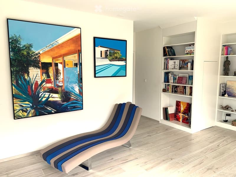 Modern room with light-colored wooden flooring, white walls, a lounge chair, built-in shelving with books and decorative items, and two large picture frames on the wall.