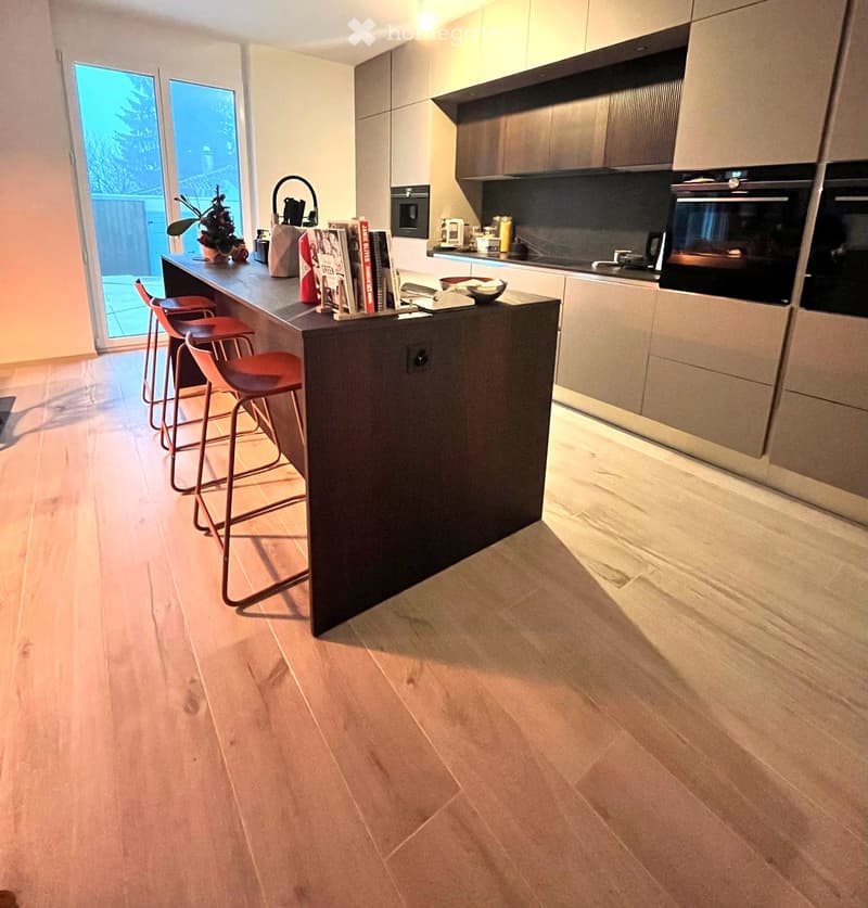 Modern kitchen with a central island, four red stools, built-in oven, and various items on the island including books, a bowl, and a plant. The kitchen features light-colored cabinets and a wooden floor.