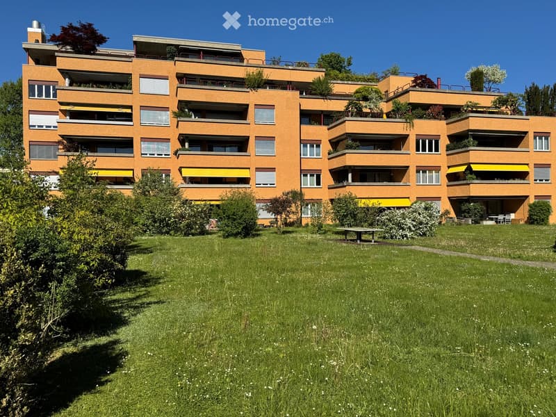 Building. Multi-story orange apartment building with balconies and greenery, surrounded by a grassy lawn and trees.