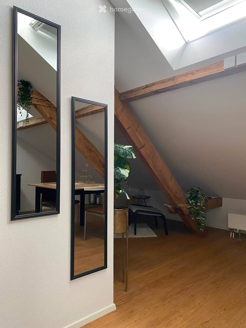 A modern, well-lit room with wooden flooring, a large mirror, a small table with chairs, a tall plant, and a sleek stool.