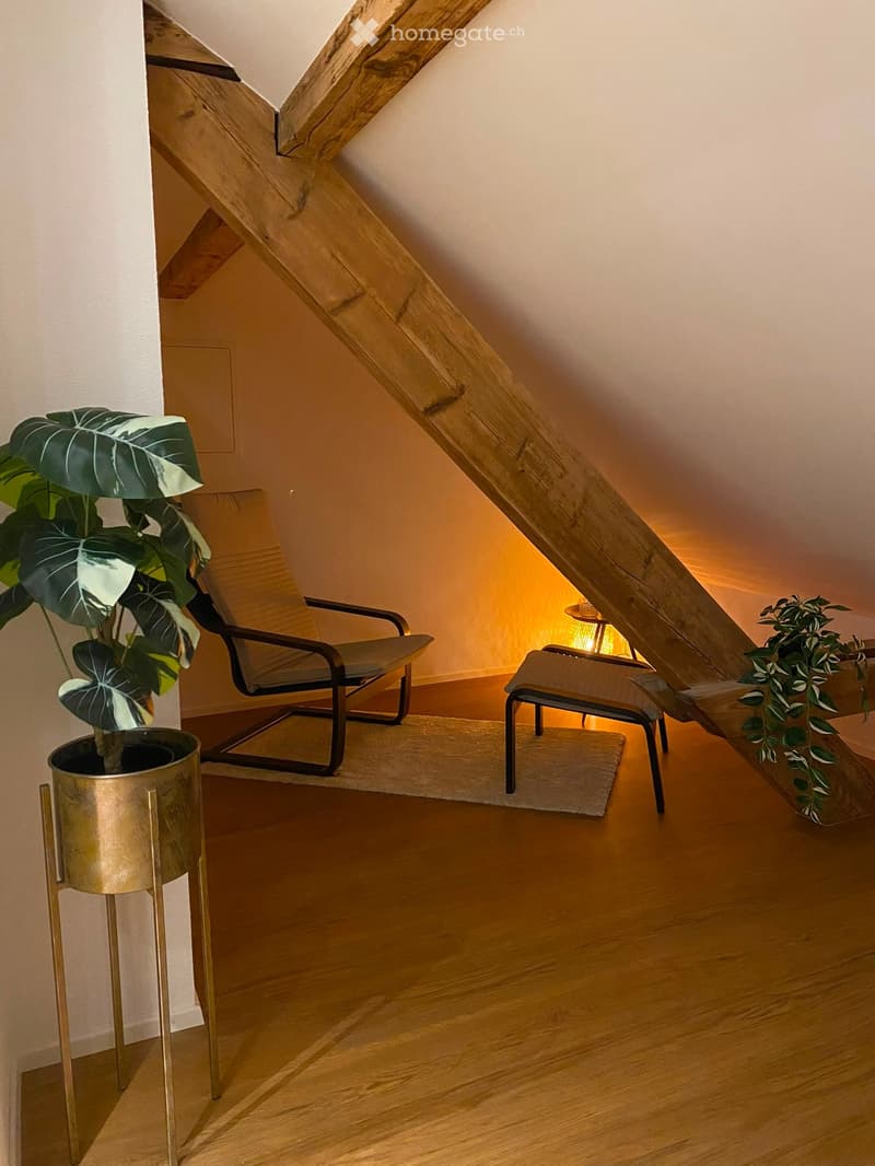 Cozy attic space with wooden floor, slanted ceiling, a chair, a small stool, a table lamp, and a large potted plant.