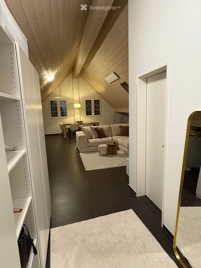 Modern living room with a sectional sofa, coffee table, and dining area in an attic space with wooden slanted ceilings and large windows.