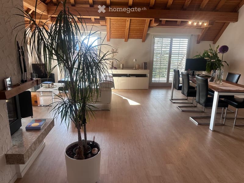 Open-plan living and dining area with wooden flooring, exposed wooden beams, large window with blinds, fireplace, sofa, coffee table, dining table with chairs, and a potted plant.