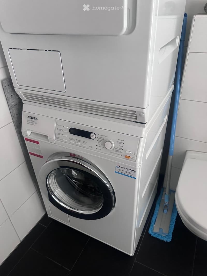 A compact bathroom with a white washing machine, a toilet, and a mop leaning against the wall.