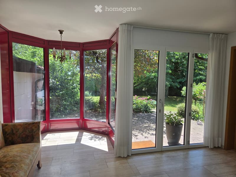 Bright living room with large windows and sliding glass doors leading to a garden, featuring a patterned sofa and a chandelier.