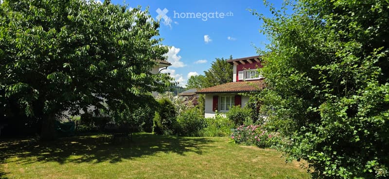 A well-maintained garden with a lawn, trees, and a small table and chairs set up under the shade of a large tree. A red house with white trim is partially visible through the foliage.