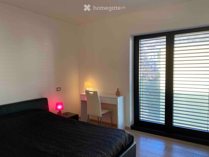 Modern bedroom with a large bed, nightstand with lamp, desk with chair, and a vase on the desk. Large windows with blinds provide natural light.