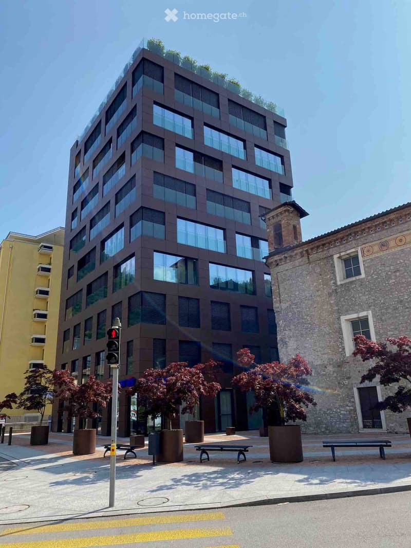 Modern multi-story building with large windows and a rooftop garden, surrounded by trees and benches in a paved area.