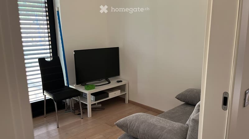 A modern living room with a grey sofa, a black chair, a flat-screen TV on a white stand, and a small table with a remote control and a green box. The room has wooden flooring and white walls.