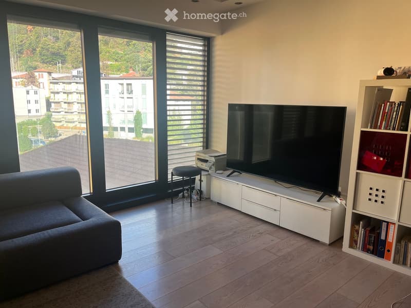 Modern living room with large windows offering a view, a grey sofa, a white TV stand with a flat-screen TV, a black stool, a printer, and a white bookcase filled with books and decorative items.