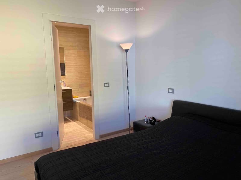 Modern bedroom with a large bed, nightstand, and floor lamp. An adjacent bathroom is visible through an open doorway, featuring a bathtub, sink, and wooden vanity.