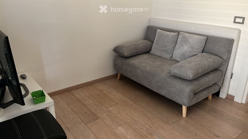 A modern living room with a grey sofa, a small white table, a screen, and a black stool. The room features light wooden flooring and white walls.