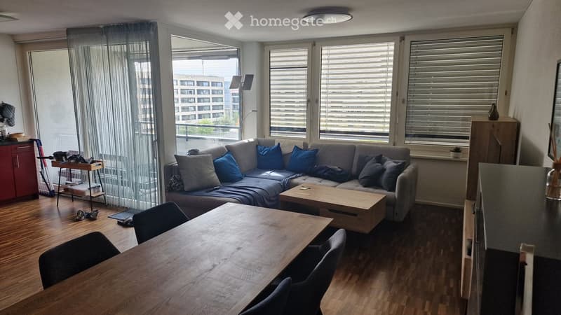 Modern living room with a grey sectional sofa, wooden coffee table, dining table with chairs, and large windows with blinds. The room features a wooden floor and a view of a cityscape.