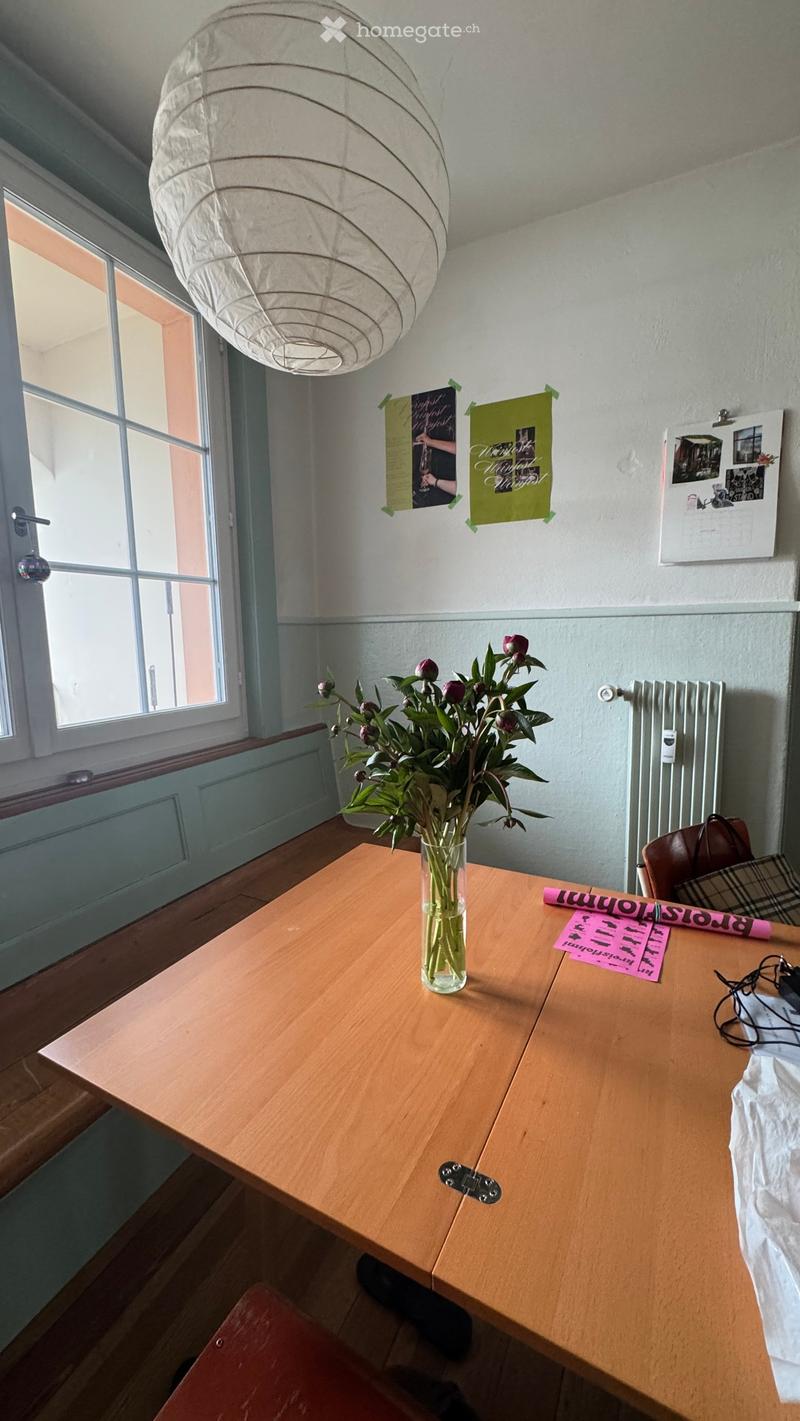 A small, well-lit room with a wooden table, vase of flowers, chair, posters, and a calendar on the wall.