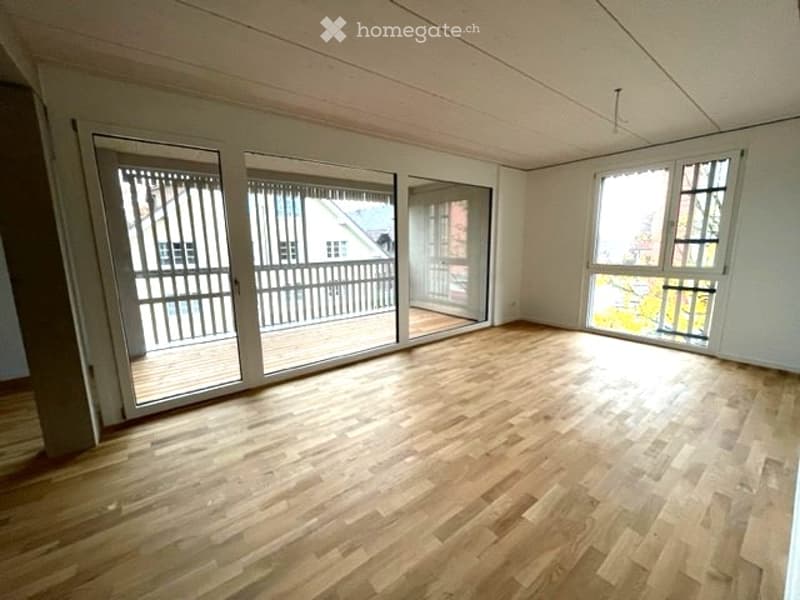 Empty room with wooden flooring, large windows, and sliding glass doors leading to a balcony.