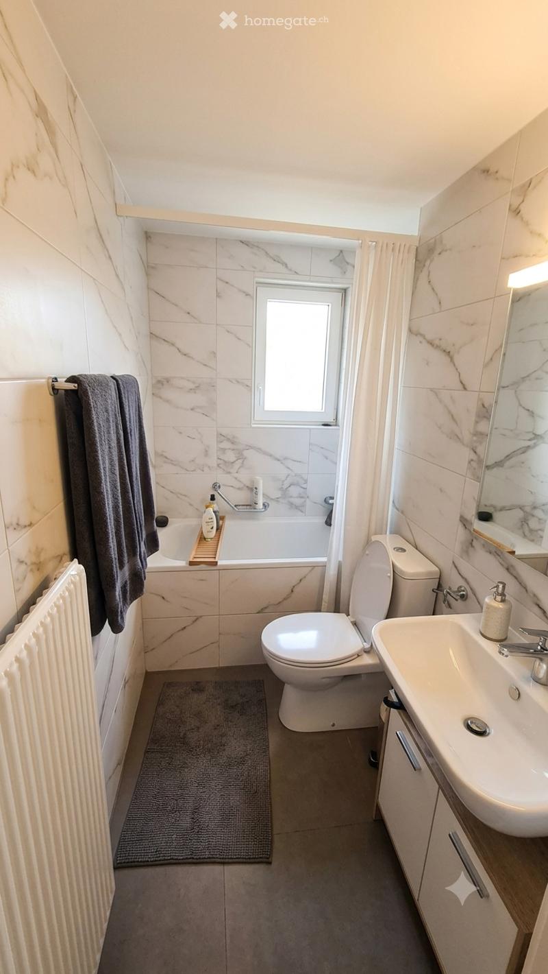 Modern bathroom with marble tiles, a bathtub, toilet, sink, mirror, and a towel on a rack. The room features a window with a curtain and a radiator on the wall.