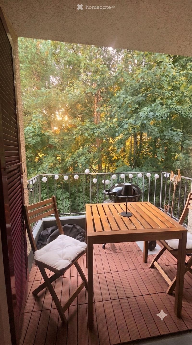 Balcony with wooden table, chairs, and a grill, surrounded by greenery.