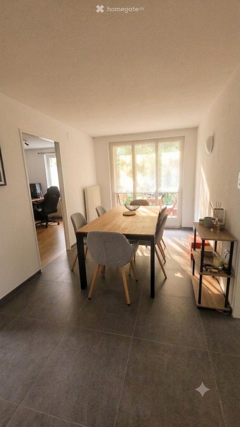 Dining room with a wooden dining table, six chairs, a small shelf, and a radiator. An office area with a chair and computer is visible in the background.