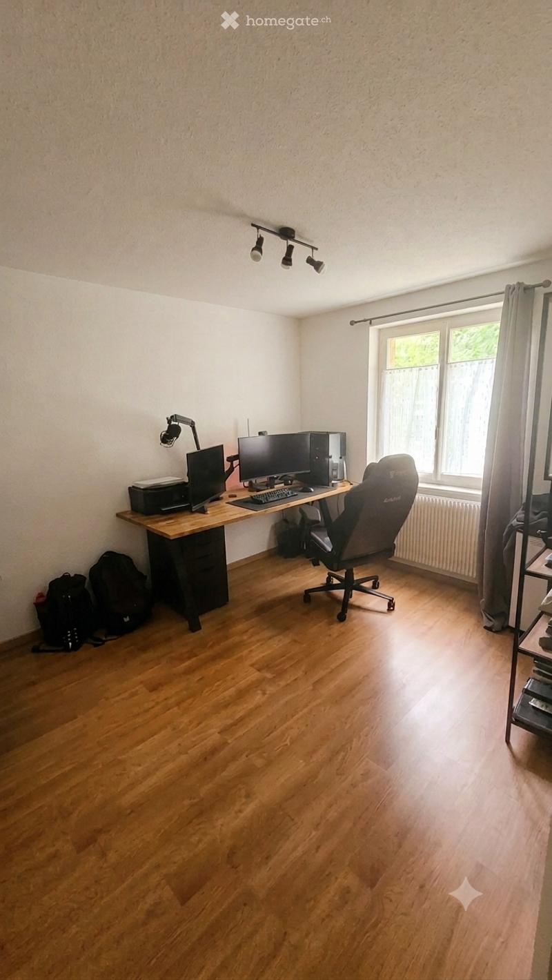 A well-lit room with wooden flooring, a large desk with dual monitors, a comfortable office chair, and a printer. Natural light streams in from a window with curtains.
