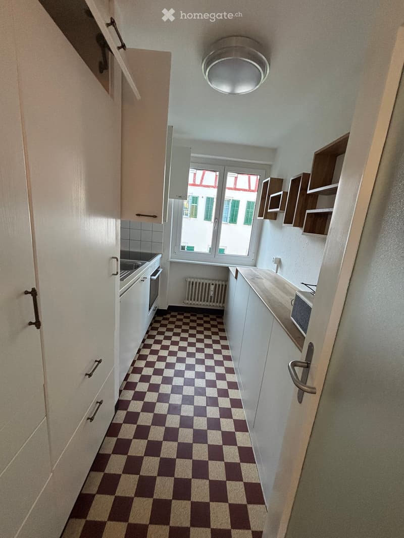 Compact kitchen with checkered floor, white cabinets, stove, oven, sink, radiator, and air conditioner. Features a window with a view of a building with green shutters.