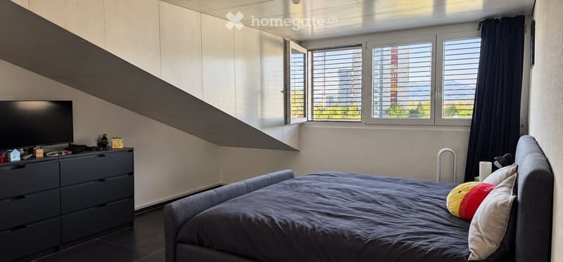 Modern bedroom with a large window offering a view, a bed with pillows, a nightstand, and a television on a dresser.