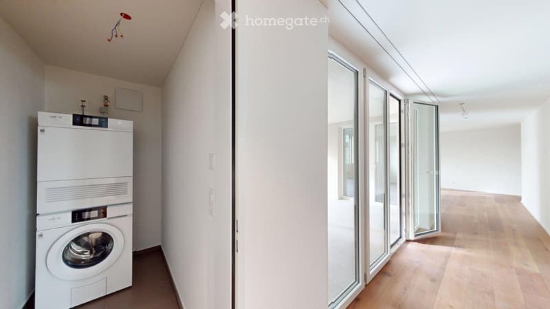 A narrow hallway with a white washing machine and dryer on the left, leading to an open space with wooden flooring and large glass doors.