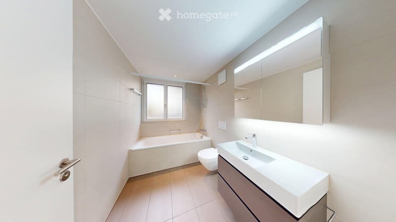 Modern bathroom with a bathtub, toilet, sink, large mirror, and built-in cabinet. Light-colored tiles and a window provide natural light.