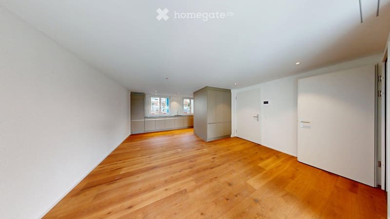 Empty room with wooden flooring, white walls, and a door leading to another room. The adjacent room features a kitchen area with cabinets and a window.