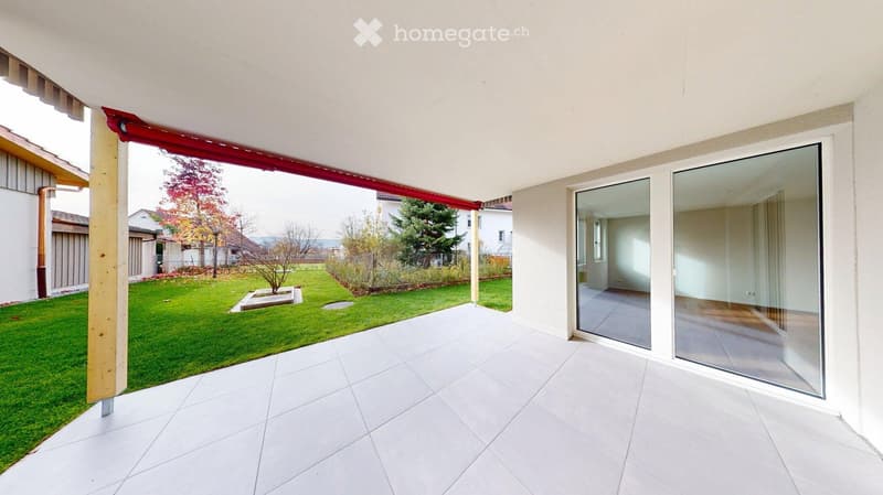 A modern, covered balcony with white tiled flooring, large glass sliding doors leading to an interior space, and a view of a well-maintained garden with green grass, trees, and a small landscaped area.