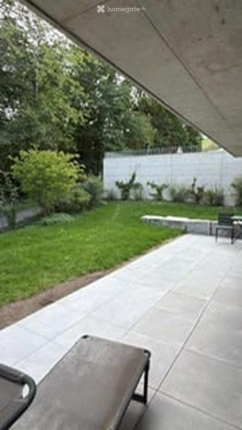 Modern patio area with concrete flooring, neatly trimmed grass, small shrubs, and a low white fence in the background. Includes a lounge chair and small table.