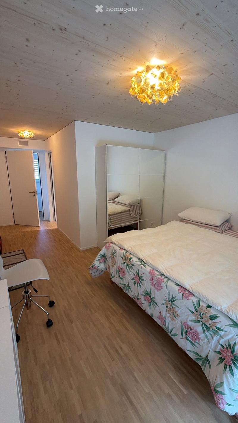 Cozy bedroom with wooden flooring, a bed with floral bedding, a white chair, a large mirror, and a wardrobe. The room features exposed wooden beams on the ceiling and warm lighting.