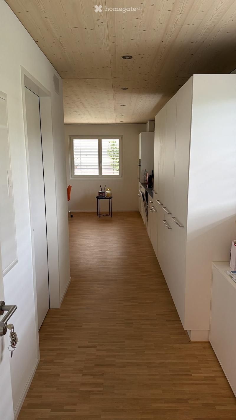 A modern, minimalist hallway with light wooden flooring, white walls, and built-in white cabinets on one side. A small round table with a few items sits near a window with blinds, and a door with a silver handle is visible on the left.