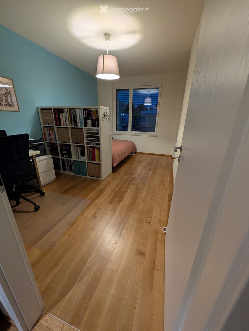 A well-lit bedroom with wooden flooring, a bed with pink bedding, a bookshelf filled with books, a desk chair, and a hanging lamp.