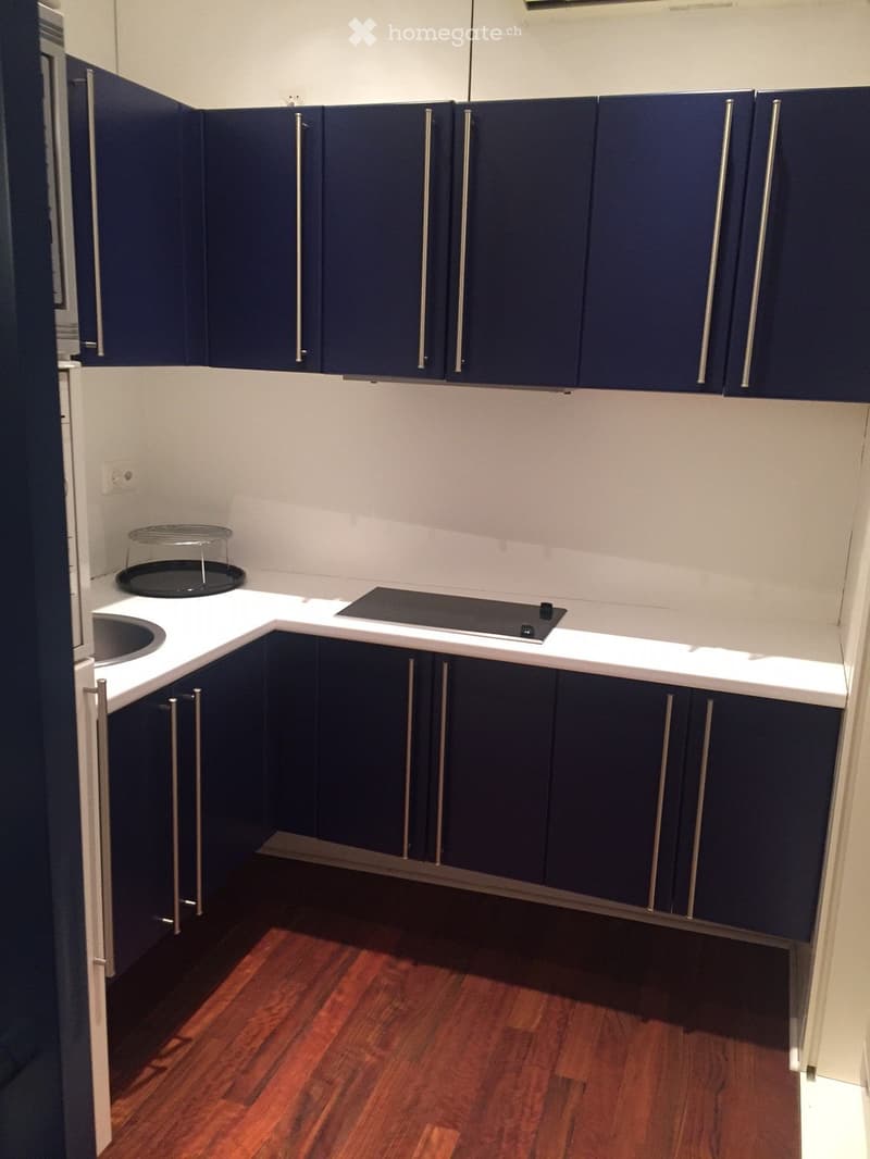Modern kitchen with dark blue cabinets, white countertops, and a wooden floor. Features a sink, stove, and a small metal basket on the counter.