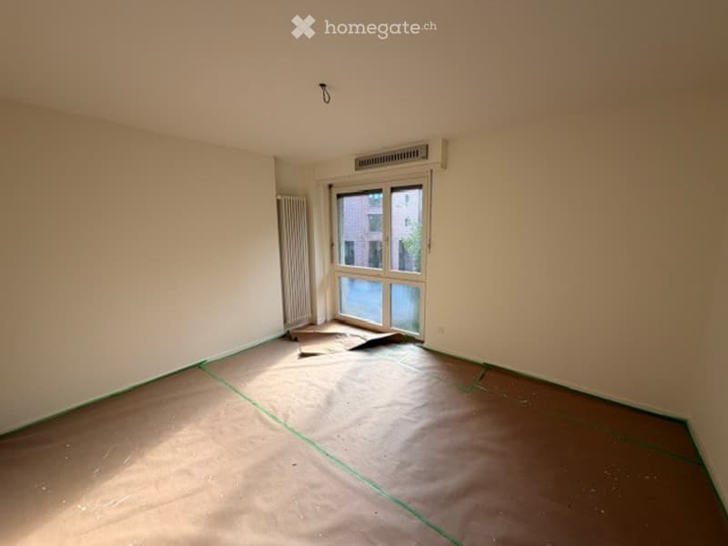 Empty room with bare walls, wooden floor, and a window with a view of a building outside. The floor is covered with protective plastic sheeting.