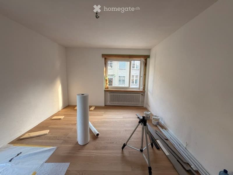 Empty room with wooden flooring, white walls, and a large window with a radiator below. Construction materials and tools are present, including a tripod, paint cans, and a roll of paper.