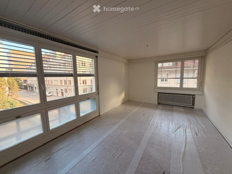 Empty room with wooden flooring, white walls, large windows with blinds, and a radiator. No furniture or equipment present.
