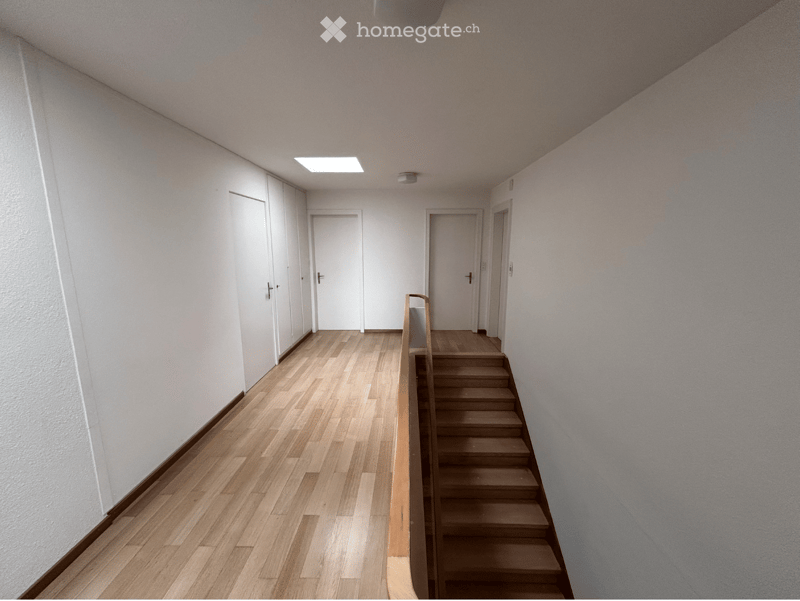 Empty hallway with wooden flooring, white walls, and a staircase leading upwards. Features include a skylight, several doors, and a handrail on the staircase.